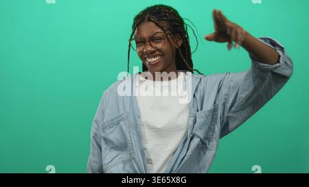 Donna che indossa occhiali e camicia in denim sorridente mentre fa il saluto con la mano in uno studio di tè; sicurezza giocosa. Foto Stock