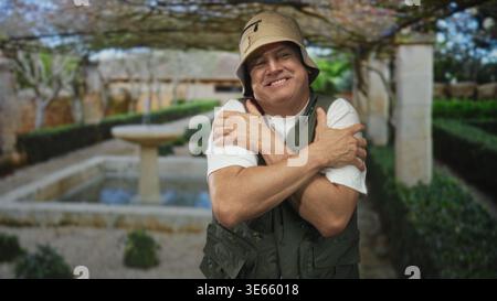 Un uomo con cappello e giubbotto da pesca attraversa le braccia che abbracciano le spalle accanto a una fontana di pietra nel cortile di un edificio, sorridendo a occhi chiusi; truffa tranquilla Foto Stock