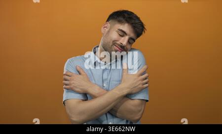 L'uomo attraversa le braccia sul petto abbracciandosi con gli occhi chiusi e un sorriso nello studio arancione; serenità. Foto Stock