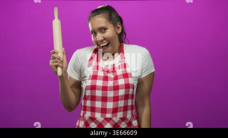 Giovane donna ispanica che cuoca tenendo in mano il mattarello e indossando un grembiule rosso a scacchi nello studio magenta; giocosa gioia di cottura. Foto Stock