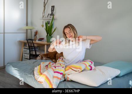 Donna felice e sognante che si sveglia al mattino soleggiato e riscalda il corpo, sedendosi nel comodo letto della camera da letto. Foto Stock