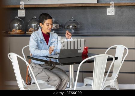 Donna afroamericana con camicia in denim seduta al bar che lavora su un computer portatile con tazza rossa e pasticcini Foto Stock