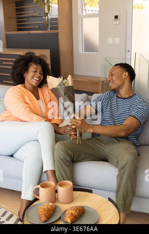 Coppia afroamericana che si scambiano bouquet avvolto mentre si siede su un divano grigio chiaro nel soggiorno Foto Stock