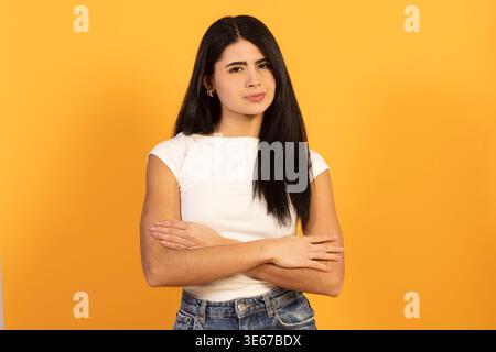 Giovane donna in piedi con le braccia incrociate, che mostra un'espressione scettica del viso e guarda direttamente la fotocamera contro un giallo luminoso Foto Stock