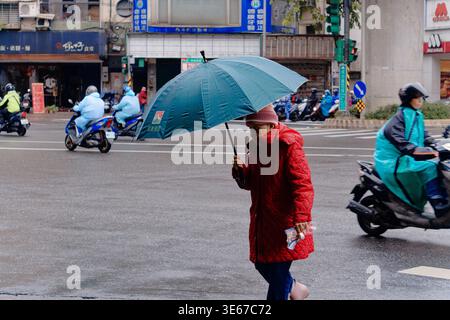 Taipei, Taiwan - 28 dicembre 2023: Una donna anziana con un cappotto rosso attraversa un incrocio piovoso di Taipei da sola, tenendo un ombrello in mezzo al traffico di scooter. Foto Stock