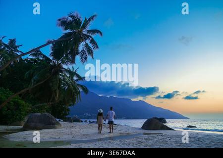 Due passeggiate mano nella mano sulla spiaggia sabbiosa di Mare Anglaise mentre il sole tramonta dietro le montagne, creando un'atmosfera romantica. Ondeggiano le palme Foto Stock