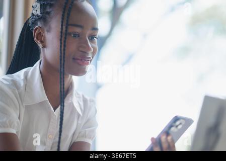 Giovane donna nera che tiene lo smartphone e guarda uno schermo in uno spazio di lavoro luminoso. Verticale laterale che mostra le comunicazioni mobili, il multitasking digitale, Foto Stock