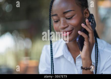 Giovane donna di colore sorridente durante una chiamata con smartphone in un bar, godendo di una comunicazione mobile amichevole. Ritratto ravvicinato con luce soffusa e vita moderna Foto Stock