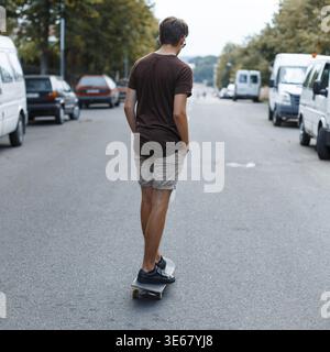Giovane uomo alla moda in abiti estivi con pantaloncini corti e canotta che cavalca uno skateboard per strada Foto Stock
