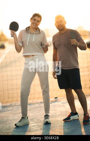 Uomo e donna sorridenti che reggono racchette da ping pong e palle su un campo al tramonto Foto Stock