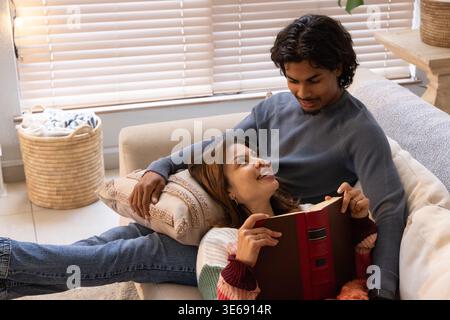 Coppia che indossa maglioni che si rilassano su un divano leggero nel soggiorno, donna che legge un libro con copertina rigida rossa Foto Stock