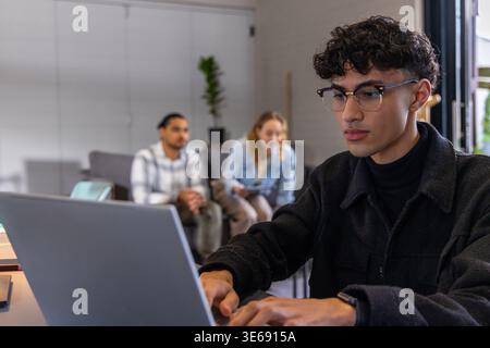 Diversi colleghi che lavorano in una moderna sala d'ufficio con un uomo asiatico che digita sul laptop vicino alla finestra Foto Stock