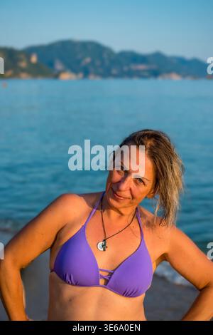 Donna felice in bikini viola che posa nel mare. Splendido turista sorridente che gode del sole estivo sulla spiaggia di Glyfada, Corfù, Grecia. Foto Stock