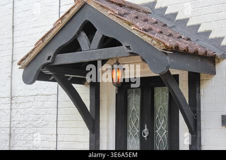 Classico portico in legno con capriate in legno scuro e tetto piastrellato, caratterizzato da una calda lanterna sospesa su una porta frontale in vetro decorativo nero. Foto Stock