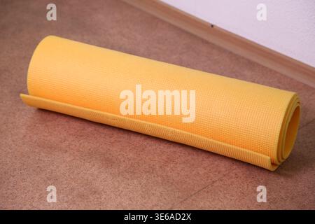 Un tappetino giallo per yoga è arrotolato e poggia su un pavimento di legno in una stanza semplice. Il tappetino è pronto per l'uso in una sessione di allenamento o in una pratica di yoga. Foto Stock