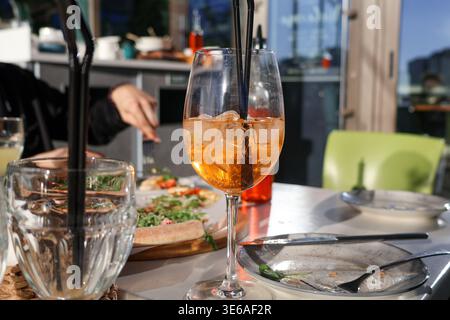 Le persone condividono un pasto in un bar con bevande sul tavolo e una vista sull'esterno durante un pomeriggio di sole. Sul tavolo sono presenti piatti e bicchieri Foto Stock