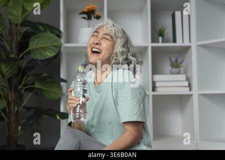 Donna anziana felice che gode di uno stile di vita sano, tiene in mano una bottiglia d'acqua e ride in un ambiente confortevole Foto Stock
