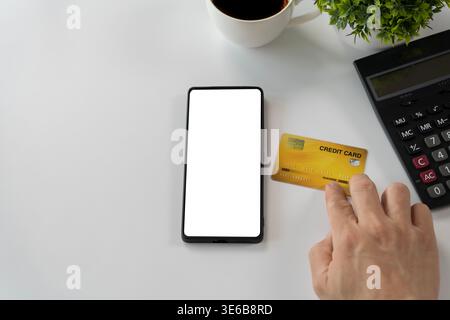 Vista dall'alto di uno smartphone vuoto accanto a una carta di credito e alla calcolatrice su una scrivania bianca pulita, adatta per pagamenti mobili, online banking, fintech, ecomme Foto Stock