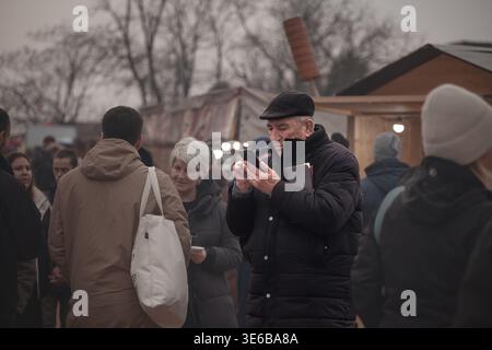 BELO BLATO, SERBIA - 7 FEBBRAIO 2026: Vecchio che usa uno smartphone nel bel mezzo di una folla a Belo Blato, Serbia, un senior specializzato nella tecnologia mobile Foto Stock