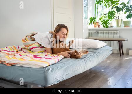 Donna positiva che si coccola con l'amato cane Vizsla in un comodo letto di casa, mostrando amore e armonia. Foto Stock