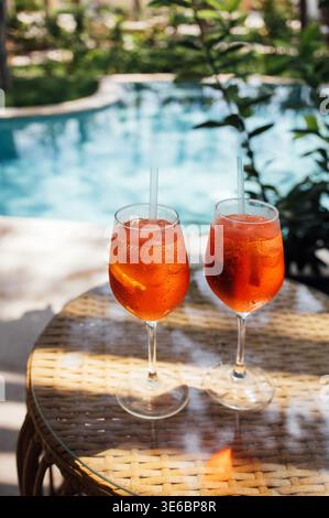 Due rinfrescanti cocktail aperol spritz in bicchieri trasparenti con ghiaccio e cannucce siedono su un tavolo di vimini accanto a una piscina con vegetazione nel retro Foto Stock