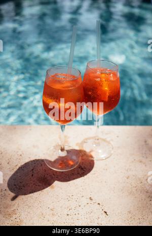 Due cocktail aperol spritz con ghiaccio e cannucce si trovano su una superficie di pietra accanto a una piscina blu chiaro, illuminata dalla luce del sole Foto Stock