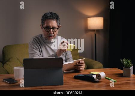 Uomo di mezza età che organizza attività personali utilizzando appunti appiccicosi mentre lavora su un tablet a casa durante la sera. Concetto di produttività, gestione del tempo, Foto Stock