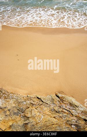 Guardando in basso l'acqua in entrata che scorre sulla spiaggia sabbiosa accanto a rocce strutturate con barnacoli bianchi Foto Stock