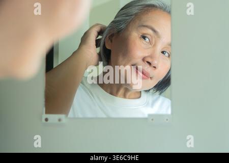 Foto di una bella donna asiatica matura di mezza età felice anziana che si coccola toccando i capelli grigi guardando se stessa allo specchio all'interno. Foto Stock