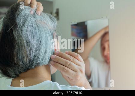 Senior attraente donna asiatica di media età 50 anni con capelli grigi che guardano riflessi a specchio pettinando capelli grigi aggrovigliati. Messa a fuoco selettiva. Foto Stock