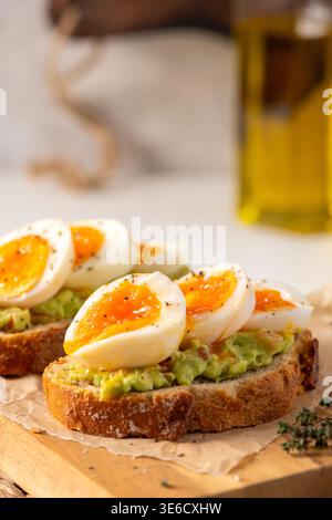 Pane artigianale croccante ricoperto di crema di avocado e uova sode di media consistenza. La luce naturale soffusa mette in risalto i tuorli stagionati e. Foto Stock