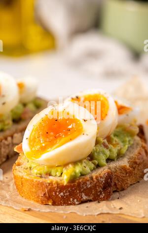 Primo piano di pane a lievitazione naturale croccante ricoperto di crema di avocado e uova sode perfettamente morbide con tuorli che cola. La scena è caratterizzata da una natura brillante Foto Stock