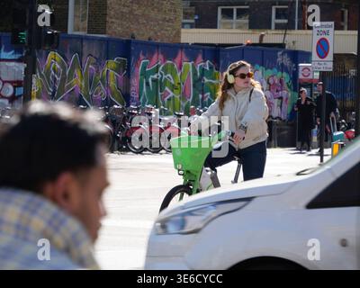 Una giovane donna che indossa cuffie e occhiali da sole aspetta su una bicicletta verde Lime a un incrocio su New Road a Whitechapel. La trafficata scena di East End Street mostra auto e pedoni che attraversano l'area vicino al Royal London Hospital e Whitechapel Road Foto Stock