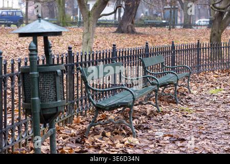 Le panchine verdi vuote del parco e il cestino in metallo sono situati tra foglie marroni d'autunno sparse, evocando un'atmosfera tranquilla e riflessiva in una città tranquilla Foto Stock