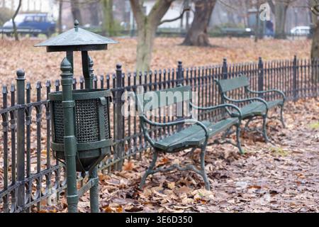 Primo piano di un cestino di metallo verde scuro con due panchine rustiche del parco dietro una recinzione ornata in ferro battuto, circondato da foglie autunnali cadute in una cal Foto Stock
