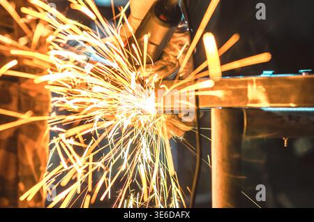 Scintille dorate derivanti dalla lucidatura dei metalli in officina. L'illuminazione intensa e lo sfondo scuro sfocato enfatizzano l'energia, il movimento e l'atmosfera industriale. Foto Stock