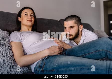 Ritratto di una donna incinta felice che tiene le scarpe per bambini e di suo marito su un divano a casa Foto Stock