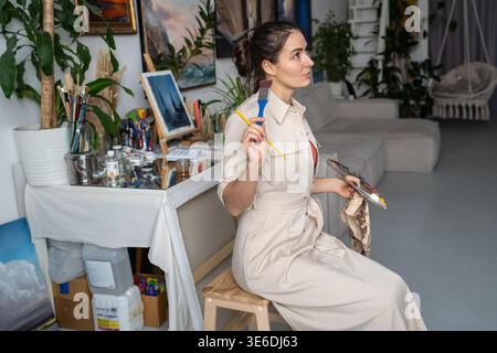 Donna abile designer artista in fase di creazione di opere d'arte, seduta a guardare dipingere su cavalletto. Foto Stock