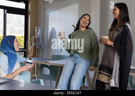 Diverse collaboratrici che chiacchierano attorno al tavolo di legno in ufficio tenendo tazze con il velo Foto Stock