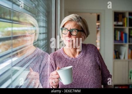 Donna anziana che prende una tazza di tè e guarda dalla finestra a casa. Foto Stock