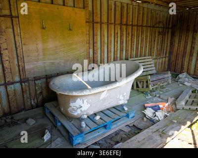 Vecchia vasca da bagno a piedi di argilla seduta su pallet di legno all'interno di un capannone fatiscente con pareti in metallo ondulato arrugginito e vari detriti sparsi, che mostrano decadimento Foto Stock