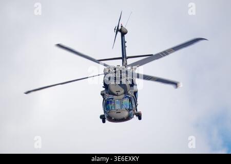 Un elicottero militare scuro con le sue lame principali del rotore sfocate in movimento vola in alto contro un cielo luminoso e nuvoloso. Foto Stock