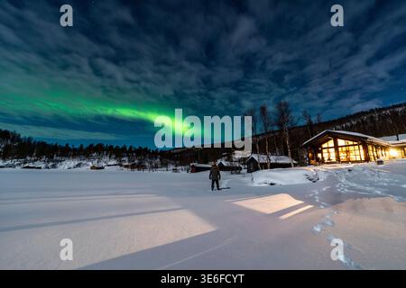 L'aurora boreale si illumina su un lago ghiacciato e su una cabina innevata vicino a Tromso, Norvegia, nella Lapponia artica. Il cielo notturno, la foresta invernale e le impronte Foto Stock
