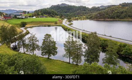 Una strada tortuosa divide due laghi nel cuore della Norvegia. Case tradizionali sorgono su una collina erbosa, incorniciata da alberi di betulla e imponenti montagne, in una Foto Stock