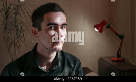 Ritratto interno di un giovane uomo che guarda la fotocamera in una stanza poco illuminata con una lampada da scrivania rossa sullo sfondo. Immagine d'atmosfera con un lato spettacolare Foto Stock