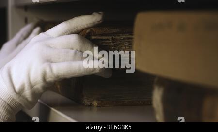 Primo piano di mano in guanto di cotone bianco che prende un libro molto vecchio dalla libreria in museo o biblioteca privata. Esplorazione degli antichi libri gialli Foto Stock