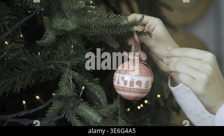 Donna che pende la palla di Natale sull'albero con le luci del bokeh. Primo piano delle mani di una donna, che sta tenendo una decorazione di Natale, una donna appeso giocattolo Foto Stock
