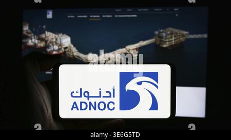 Stoccarda, Germania - 12-08-2024: Persona che detiene un cellulare con il logo dell'azienda petrolifera Abu Dhabi National Oil Company (ADNOC) di fronte alle pagine web aziendali Foto Stock