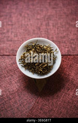 Un vivace assortimento di foglie di tè alle erbe essiccate disposte in una ciotola bianca su un ricco sfondo rosso Foto Stock