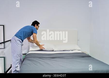 Uomo che organizza biancheria da letto e cuscini, esegue le pulizie di routine in camera da letto. Concentrarsi sulla vita domestica e sull'organizzazione Foto Stock